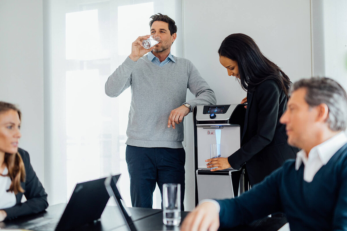 Büro in dem sich eine Frau Wasser aus dem Waterlogic Wasserspender holt während ein Mann dagegen lehnt und Wasser trinkt 