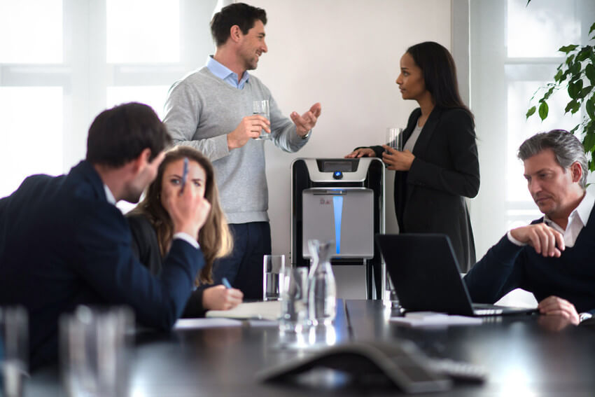 Meetingraum mit Personen im Gespräch vertieft und eine Frau holt sich Wasser aus einem Waterlogic Wasserspender während Sie sich mit einem Mann unterhält  